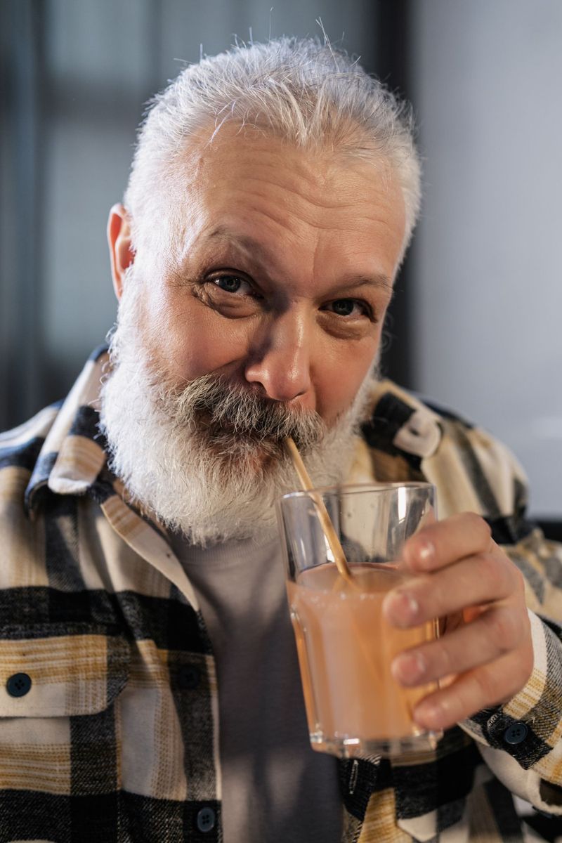 Äldre man med grått skägg som dricker en smoothie med sugrör